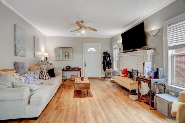 living area featuring crown molding, light wood-style flooring, a brick fireplace, ceiling fan, and a textured ceiling