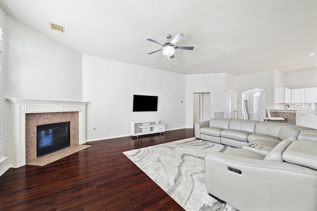 living room featuring visible vents, arched walkways, a ceiling fan, a tiled fireplace, and dark wood-style floors