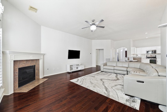 living area featuring arched walkways, dark wood finished floors, visible vents, a tiled fireplace, and a ceiling fan