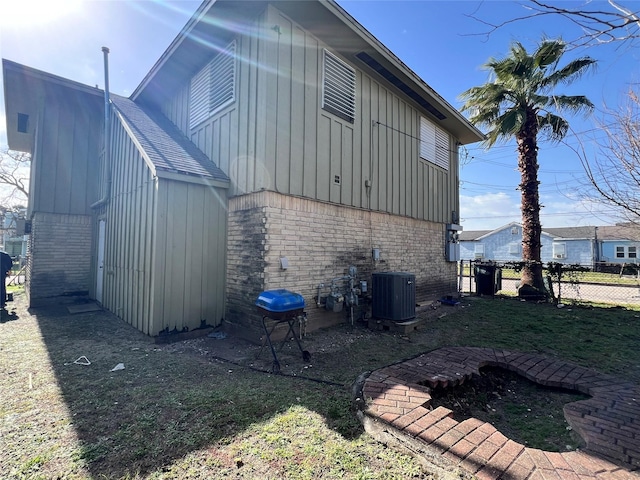 view of side of home with brick siding, a lawn, central AC unit, board and batten siding, and fence