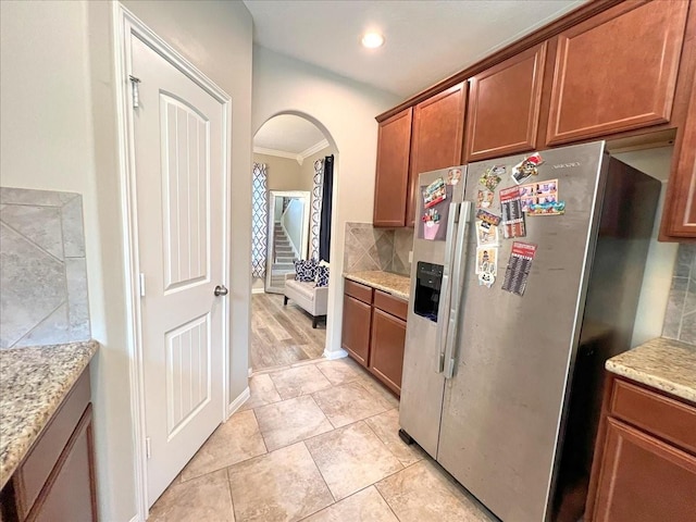 kitchen featuring arched walkways, light stone countertops, backsplash, brown cabinets, and stainless steel fridge