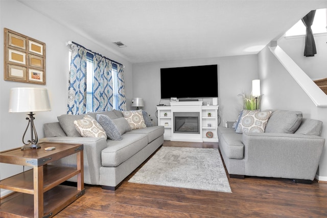 living area featuring stairs, a glass covered fireplace, dark wood finished floors, and visible vents