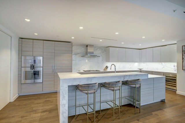 kitchen with stainless steel double oven, modern cabinets, light wood-style floors, and wall chimney range hood