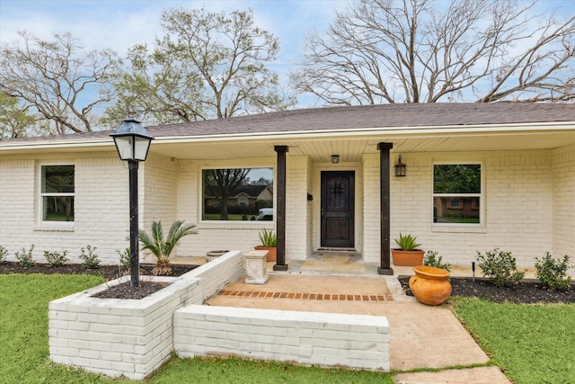 view of exterior entry featuring a yard, brick siding, roof with shingles, and a porch