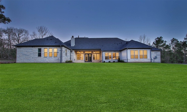 back of house at dusk featuring a shingled roof, french doors, stone siding, a chimney, and a yard