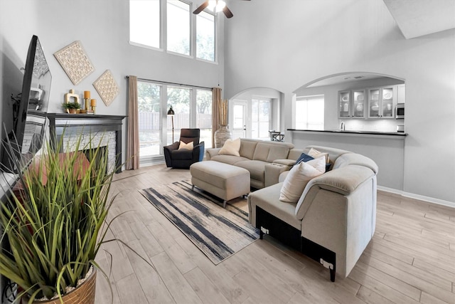 living area featuring baseboards, ceiling fan, a high ceiling, a stone fireplace, and light wood-style floors