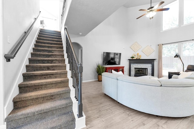 living area featuring arched walkways, stairway, a ceiling fan, a stone fireplace, and light wood-type flooring