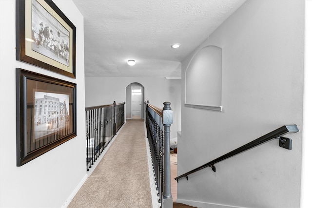 hallway featuring a textured ceiling, arched walkways, light carpet, an upstairs landing, and baseboards