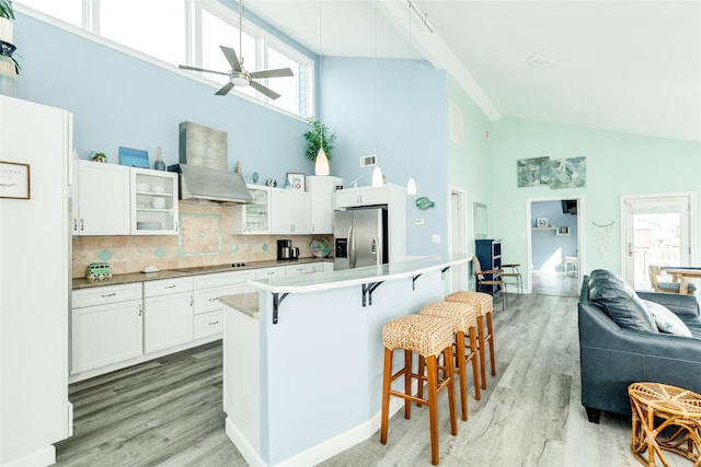 kitchen featuring black electric stovetop, a kitchen breakfast bar, white cabinets, wall chimney exhaust hood, and stainless steel fridge