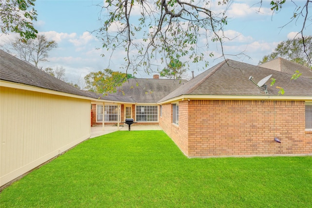 rear view of property with a shingled roof, brick siding, a lawn, and a patio