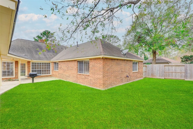 rear view of property with roof with shingles, a yard, brick siding, a patio, and fence