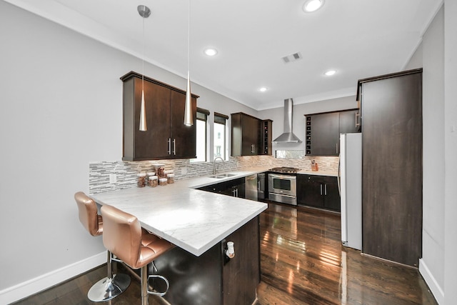 kitchen with a peninsula, a breakfast bar, stainless steel appliances, hanging light fixtures, and wall chimney range hood