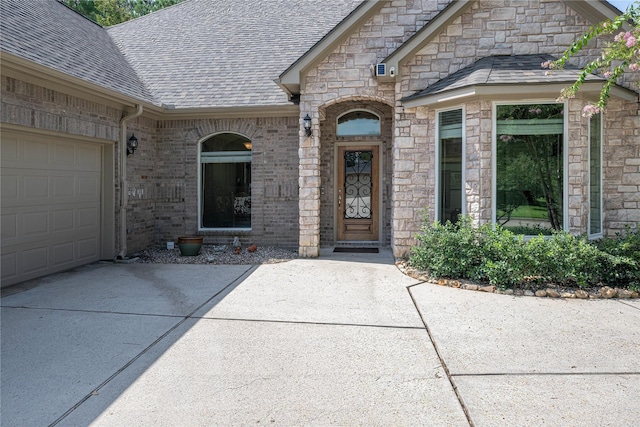 view of exterior entry with an attached garage, driveway, a shingled roof, and brick siding