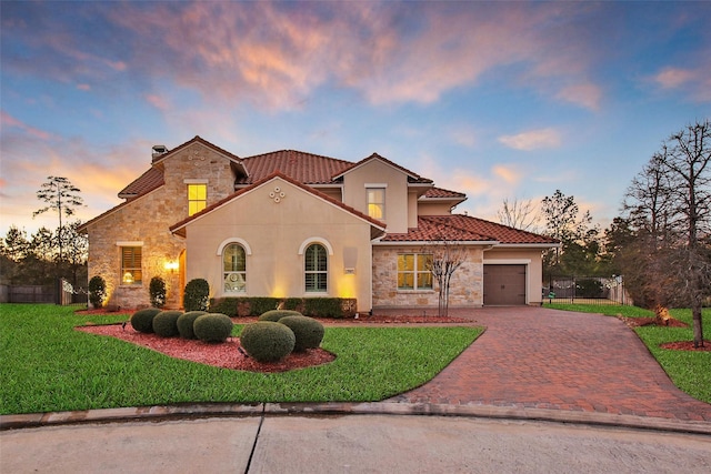 mediterranean / spanish-style home featuring an attached garage, stone siding, decorative driveway, a front lawn, and a chimney