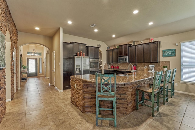 kitchen with arched walkways, dark brown cabinetry, stainless steel appliances, a breakfast bar, and dark stone counters