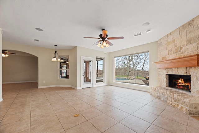 unfurnished living room with light tile patterned floors, visible vents, arched walkways, ceiling fan, and a stone fireplace