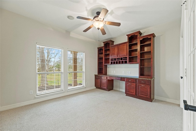 unfurnished office with light carpet, built in desk, a ceiling fan, and baseboards