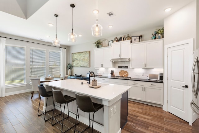 kitchen with dark countertops, an island with sink, white cabinetry, pendant lighting, and backsplash