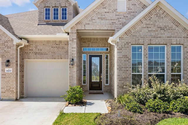 view of exterior entry featuring a garage, driveway, brick siding, and roof with shingles