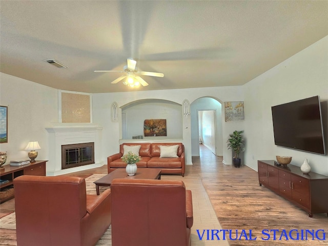 living room with arched walkways, ceiling fan, wood finished floors, visible vents, and a glass covered fireplace