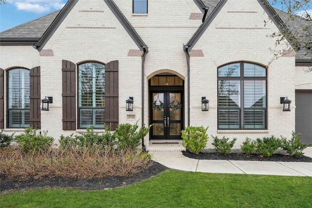 view of exterior entry featuring french doors, roof with shingles, and brick siding