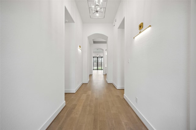 corridor featuring arched walkways, a chandelier, light wood finished floors, and baseboards