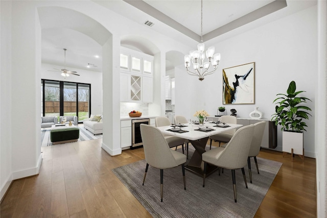 dining area featuring wood finished floors, ceiling fan with notable chandelier, beverage cooler, arched walkways, and visible vents