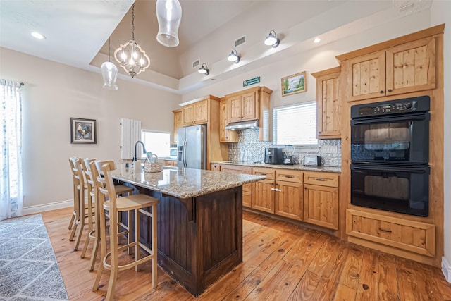 kitchen with an island with sink, light stone counters, hanging light fixtures, under cabinet range hood, and black appliances