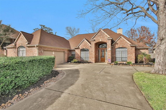 french country inspired facade featuring brick siding, roof with shingles, a chimney, a garage, and driveway