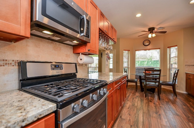 kitchen with appliances with stainless steel finishes, brown cabinets, dark wood-style flooring, light stone countertops, and backsplash