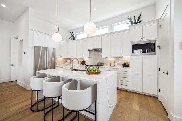 kitchen featuring an island with sink, under cabinet range hood, stainless steel appliances, backsplash, and a sink