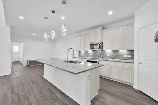 kitchen featuring stainless steel microwave, a sink, decorative backsplash, an island with sink, and dark wood finished floors