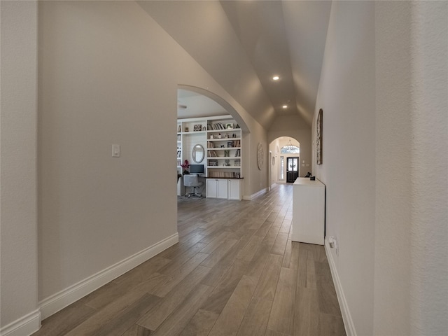 hall with built in shelves, wood finished floors, baseboards, lofted ceiling, and arched walkways