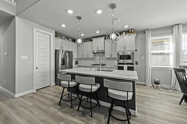 kitchen with decorative light fixtures, stainless steel appliances, light countertops, white cabinetry, and an island with sink