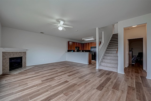 unfurnished living room featuring visible vents, a tiled fireplace, ceiling fan, stairs, and light wood-type flooring