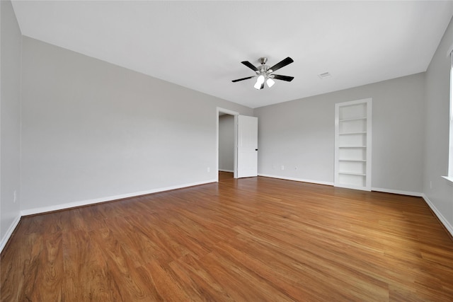 empty room featuring ceiling fan, built in shelves, wood finished floors, and baseboards