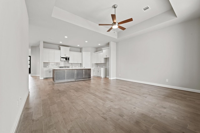 unfurnished living room featuring a tray ceiling, visible vents, light wood-style flooring, a ceiling fan, and baseboards