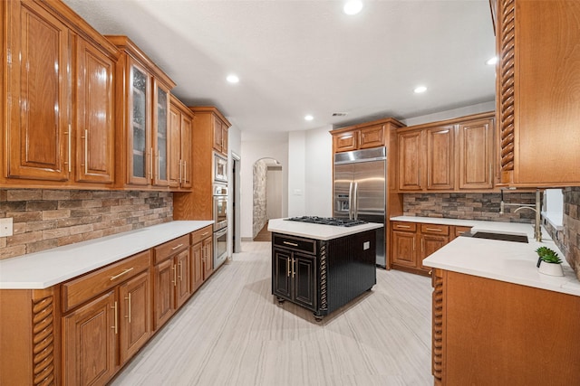 kitchen with built in appliances, arched walkways, a sink, brown cabinetry, and glass insert cabinets