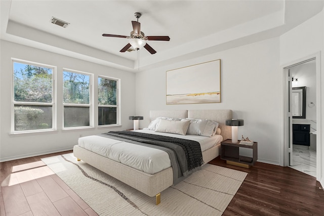 bedroom with baseboards, a raised ceiling, wood finished floors, and ensuite bathroom