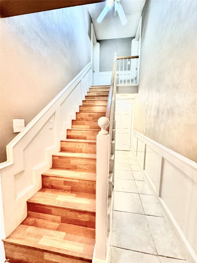 staircase featuring wainscoting, tile patterned flooring, a ceiling fan, and a decorative wall