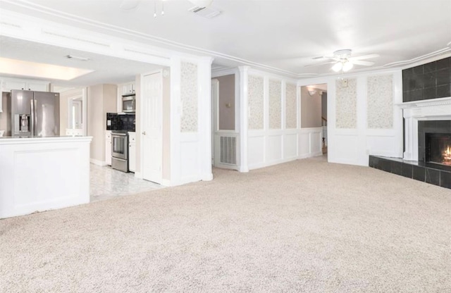 unfurnished living room featuring a tiled fireplace, a decorative wall, a ceiling fan, and light colored carpet