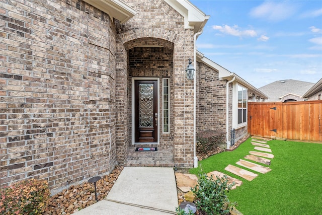 view of exterior entry with a yard, brick siding, and fence