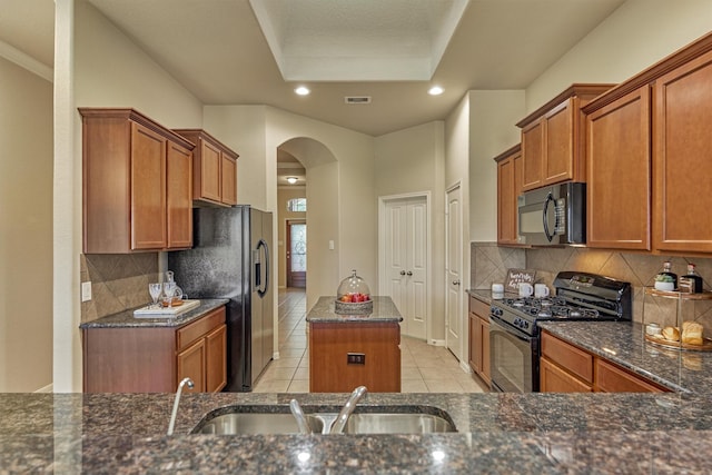 kitchen with arched walkways, dark stone counters, brown cabinetry, black appliances, and a sink