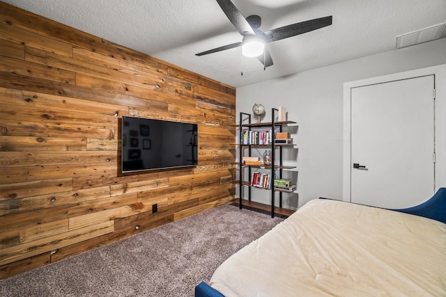 carpeted bedroom with a textured ceiling, wood walls, visible vents, and a ceiling fan