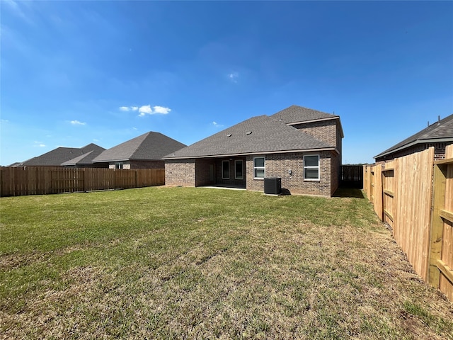 rear view of property with central AC unit, a fenced backyard, roof with shingles, a yard, and brick siding