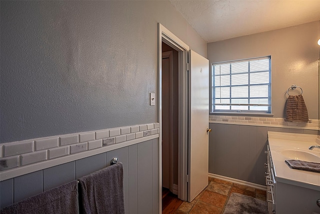 bathroom featuring a textured ceiling, stone tile floors, wood walls, vanity, and wainscoting