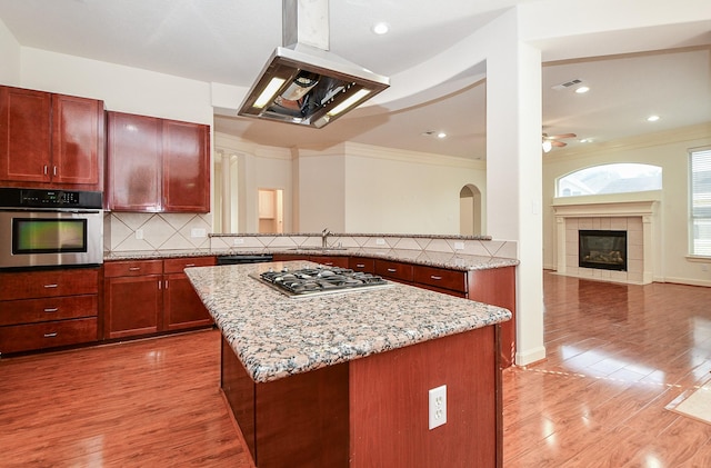 kitchen with light stone countertops, island range hood, stainless steel appliances, light wood-style floors, and decorative backsplash