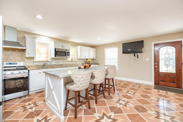 kitchen with a kitchen bar, a sink, stainless steel appliances, wall chimney range hood, and baseboards