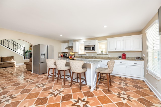 kitchen featuring a kitchen island, wall chimney range hood, a kitchen breakfast bar, appliances with stainless steel finishes, and white cabinets