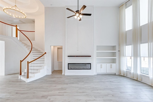 unfurnished living room featuring light wood-type flooring, stairs, a towering ceiling, and a glass covered fireplace